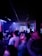 A lively nightclub scene with a crowd of people dancing and enjoying themselves under dim lighting. A DJ is positioned on a raised platform in the background, surrounded by equipment and bright spotlights casting vibrant colors.