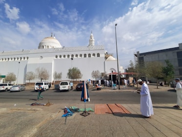 A white mosque with domes and a minaret is the central structure. Several people are gathered outside, some are setting up or selling items on the sidewalk. There are trees and parked cars in front of the mosque, and the sky is partly cloudy.
