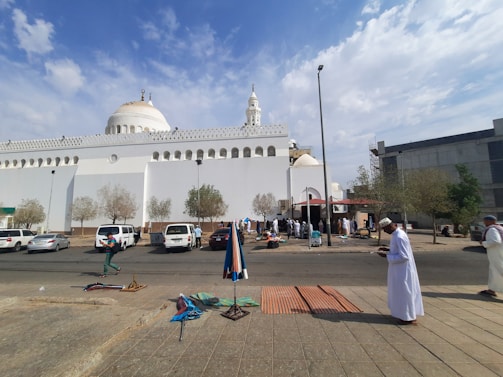A white mosque with domes and a minaret is the central structure. Several people are gathered outside, some are setting up or selling items on the sidewalk. There are trees and parked cars in front of the mosque, and the sky is partly cloudy.