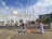A white mosque with domes and a minaret is the central structure. Several people are gathered outside, some are setting up or selling items on the sidewalk. There are trees and parked cars in front of the mosque, and the sky is partly cloudy.
