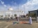 A white mosque with domes and a minaret is the central structure. Several people are gathered outside, some are setting up or selling items on the sidewalk. There are trees and parked cars in front of the mosque, and the sky is partly cloudy.