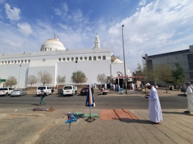 A white mosque with domes and a minaret is the central structure. Several people are gathered outside, some are setting up or selling items on the sidewalk. There are trees and parked cars in front of the mosque, and the sky is partly cloudy.
