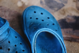 A pair of blue rubber clogs with perforations on the upper surface is placed on a textured surface. The shoes display a slightly worn appearance with some dirt and markings visible, adding a rustic feel to the overall scene.