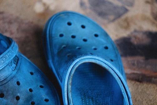 A pair of blue rubber clogs with perforations on the upper surface is placed on a textured surface. The shoes display a slightly worn appearance with some dirt and markings visible, adding a rustic feel to the overall scene.