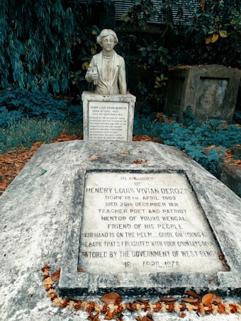 A statue of Henry Louis Vivian Derozio stands as a central feature, surrounded by dense foliage. In front of the statue is a gravestone with inscriptions commemorating Derozio as a teacher, poet, and prominent figure in the Bengal Renaissance. The area is adorned with fallen leaves and dried flowers, adding a sense of age and reverence to the scene.