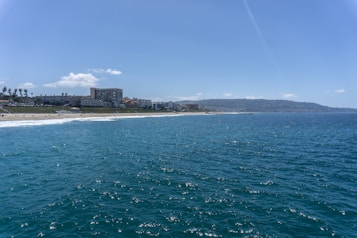 A vast ocean stretches towards the horizon with gentle waves. The coastline is bordered by sandy beaches and a series of buildings, including hotels or apartments. Sparse palm trees dot the landscape, and hills rise in the distance beneath a clear blue sky dotted with a few clouds.