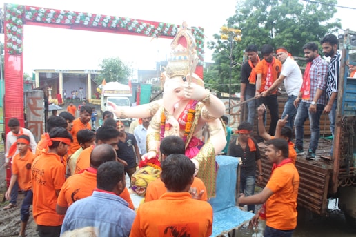 A large sculpture of a deity, Ganesha, surrounded by a group of people. Most of the people are dressed in orange, some wearing headbands, and appear to be participating in a festival or procession. The sculpture is richly decorated with floral garlands. In the background, there are decorative arches and some greenery, indicating an outdoor setting.