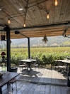 A cozy outdoor dining area with wooden flooring and an overhead wooden roof adorned with hanging lights, overlooking a lush vineyard. The dining tables are set for guests, surrounded by greenery and a scenic view of distant hills under a clear sky.