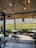 A cozy outdoor dining area with wooden flooring and an overhead wooden roof adorned with hanging lights, overlooking a lush vineyard. The dining tables are set for guests, surrounded by greenery and a scenic view of distant hills under a clear sky.