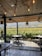 A cozy outdoor dining area with wooden flooring and an overhead wooden roof adorned with hanging lights, overlooking a lush vineyard. The dining tables are set for guests, surrounded by greenery and a scenic view of distant hills under a clear sky.