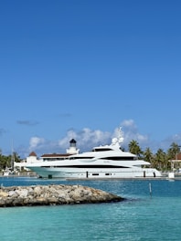 A large luxury yacht docked at a marina with a lighthouse and palm trees in the background under a clear blue sky. The water is a vibrant turquoise color and a rocky pier extends into the sea.