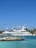 A large luxury yacht docked at a marina with a lighthouse and palm trees in the background under a clear blue sky. The water is a vibrant turquoise color and a rocky pier extends into the sea.