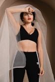 A woman is posing under a sheer, translucent fabric draped over her. She is wearing a black sleeveless top and high-waisted black Adidas leggings with white stripes. Her arm is raised, resting on her forehead, and she is adorned with large, gold hoop earrings. The background has a soft, neutral tone.