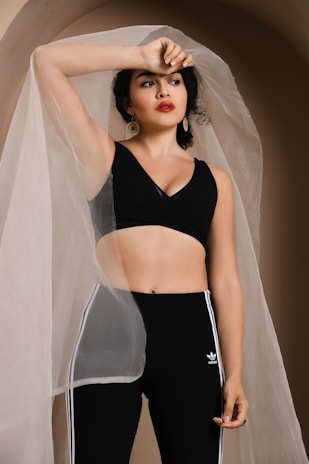 A woman is posing under a sheer, translucent fabric draped over her. She is wearing a black sleeveless top and high-waisted black Adidas leggings with white stripes. Her arm is raised, resting on her forehead, and she is adorned with large, gold hoop earrings. The background has a soft, neutral tone.