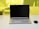 A laptop with a black screen is placed on a white desk. Next to it, on the right side, is a black wireless mouse. The background shows a yellow-tinted glass partition with a blurred view of some objects including a pen holder and another item.