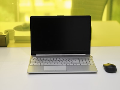 A laptop with a black screen is placed on a white desk. Next to it, on the right side, is a black wireless mouse. The background shows a yellow-tinted glass partition with a blurred view of some objects including a pen holder and another item.