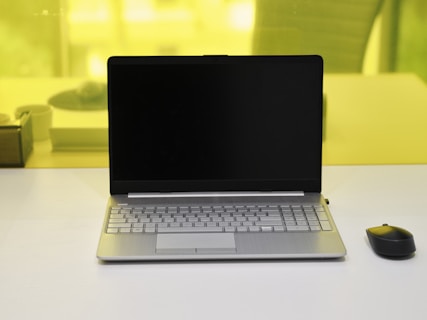 A laptop with a black screen is placed on a white desk. Next to it, on the right side, is a black wireless mouse. The background shows a yellow-tinted glass partition with a blurred view of some objects including a pen holder and another item.