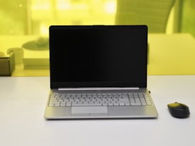 A laptop with a black screen is placed on a white desk. Next to it, on the right side, is a black wireless mouse. The background shows a yellow-tinted glass partition with a blurred view of some objects including a pen holder and another item.