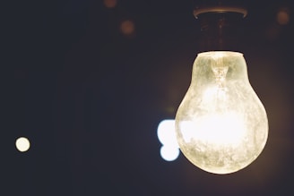 A glowing light bulb is prominently displayed against a dark background, illuminating its immediate surroundings with a warm glow. The glass of the bulb is slightly dusty, adding texture to the scene. Dim lights or reflections appear in the background, adding depth and contrast.