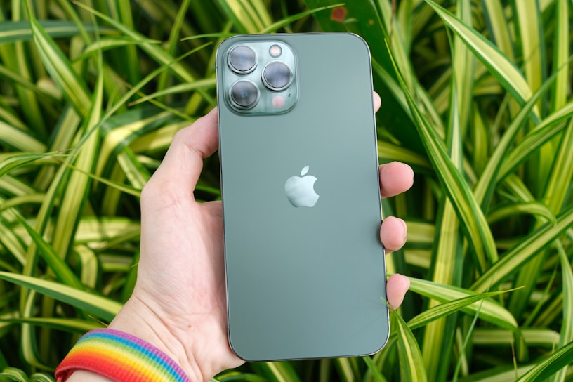 A hand holding a green smartphone with a visible Apple logo on its back, set against a backdrop of lush green plants with variegated leaves. The phone's camera lenses are prominent, and the person is wearing a colorful, rainbow-striped wristband.