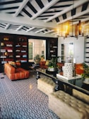 An elegant and stylish living room features a geometric patterned floor, leather sofas, and a central black table. Bookshelves line one wall, while a modern chandelier illuminates the space. Decorative plants and candles add a touch of nature and coziness.
