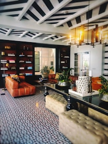 An elegant and stylish living room features a geometric patterned floor, leather sofas, and a central black table. Bookshelves line one wall, while a modern chandelier illuminates the space. Decorative plants and candles add a touch of nature and coziness.