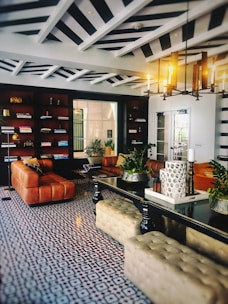An elegant and stylish living room features a geometric patterned floor, leather sofas, and a central black table. Bookshelves line one wall, while a modern chandelier illuminates the space. Decorative plants and candles add a touch of nature and coziness.