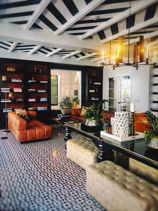 An elegant and stylish living room features a geometric patterned floor, leather sofas, and a central black table. Bookshelves line one wall, while a modern chandelier illuminates the space. Decorative plants and candles add a touch of nature and coziness.