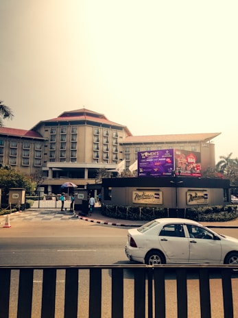 A large hotel building with multiple windows and a red rooftop is visible in the background. In front of the hotel, there is a billboard with advertisements. A car is parked on the road in front of the hotel entrance, where a couple of people are standing near the gate, one holding an umbrella.