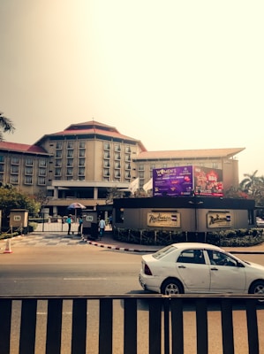 A large hotel building with multiple windows and a red rooftop is visible in the background. In front of the hotel, there is a billboard with advertisements. A car is parked on the road in front of the hotel entrance, where a couple of people are standing near the gate, one holding an umbrella.
