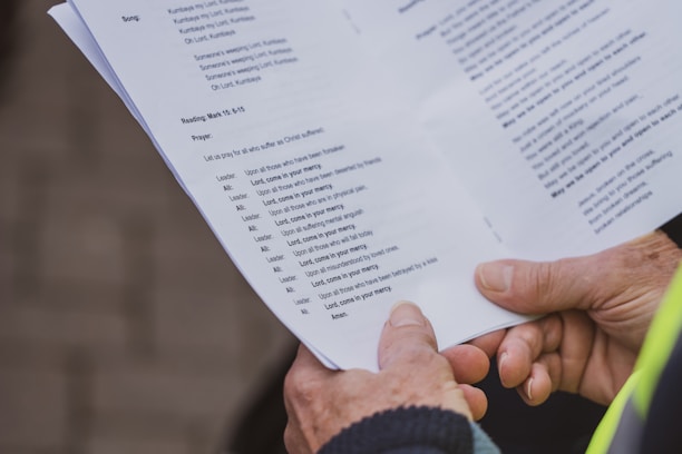 A person is holding a booklet with printed text, possibly religious content, including prayers and readings. The text is clear and the booklet is held in both hands.