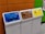 Three recycling bins are lined up next to each other against a colorful wall. The bins are labeled for different materials: paper with a blue top, plastic with a brown top, and aluminum with a yellow top. Each bin has a distinct shape for the deposit slot appropriate for the material.