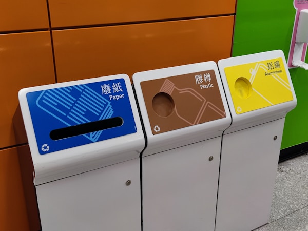 Three recycling bins are lined up next to each other against a colorful wall. The bins are labeled for different materials: paper with a blue top, plastic with a brown top, and aluminum with a yellow top. Each bin has a distinct shape for the deposit slot appropriate for the material.