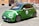 A green compact car is parked on an urban street. The car has a sporty appearance with racing stripes and the number 4 displayed on the side. It is set against a backdrop of European-style buildings with a warm tone.