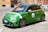 A green compact car is parked on an urban street. The car has a sporty appearance with racing stripes and the number 4 displayed on the side. It is set against a backdrop of European-style buildings with a warm tone.