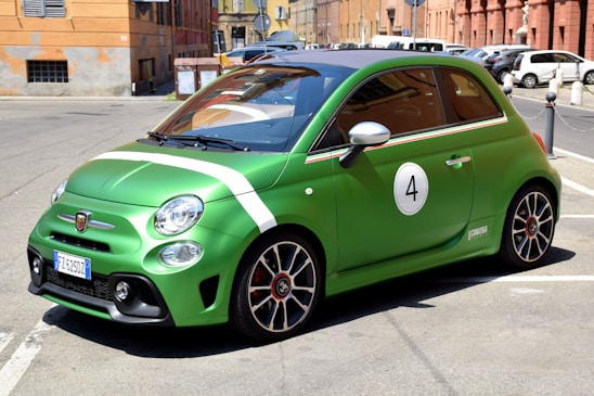 A green compact car is parked on an urban street. The car has a sporty appearance with racing stripes and the number 4 displayed on the side. It is set against a backdrop of European-style buildings with a warm tone.