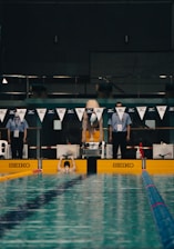 A swimmer is performing a backstroke start at a competitive swimming pool. There are officials standing on either side of the starting block, and the pool is marked with colorful lane dividers. The setting appears to be indoors with subdued lighting.