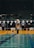A swimmer is performing a backstroke start at a competitive swimming pool. There are officials standing on either side of the starting block, and the pool is marked with colorful lane dividers. The setting appears to be indoors with subdued lighting.