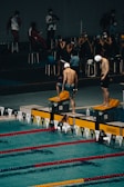 Swimmers are preparing for a race at an indoor swimming pool. The scene captures two swimmers in swim caps and shorts standing on starting blocks at the edge of the pool, with others sitting and observing from the background. The swimming lanes are marked with red and yellow lane dividers.