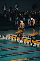 Swimmers are preparing for a race at an indoor swimming pool. The scene captures two swimmers in swim caps and shorts standing on starting blocks at the edge of the pool, with others sitting and observing from the background. The swimming lanes are marked with red and yellow lane dividers.