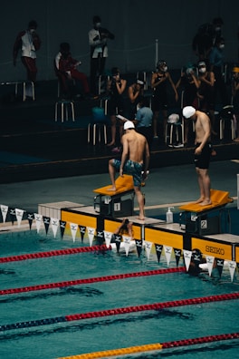 Swimmers are preparing for a race at an indoor swimming pool. The scene captures two swimmers in swim caps and shorts standing on starting blocks at the edge of the pool, with others sitting and observing from the background. The swimming lanes are marked with red and yellow lane dividers.