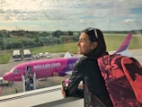 A woman with short hair, wearing sunglasses on her head and a black leather jacket, stands inside an airport terminal, gazing out the window. She carries a pink and black backpack and holds a smartphone. Outside, a pink Wizz Air airplane is visible with passengers boarding it via a movable staircase. The background features a lush landscape with trees and a partly cloudy sky.