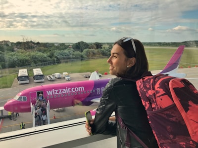 A woman with short hair, wearing sunglasses on her head and a black leather jacket, stands inside an airport terminal, gazing out the window. She carries a pink and black backpack and holds a smartphone. Outside, a pink Wizz Air airplane is visible with passengers boarding it via a movable staircase. The background features a lush landscape with trees and a partly cloudy sky.