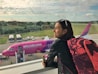 A woman with short hair, wearing sunglasses on her head and a black leather jacket, stands inside an airport terminal, gazing out the window. She carries a pink and black backpack and holds a smartphone. Outside, a pink Wizz Air airplane is visible with passengers boarding it via a movable staircase. The background features a lush landscape with trees and a partly cloudy sky.