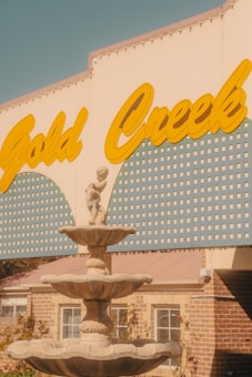 A vintage-style sign reading 'Gold Creek' in bold yellow letters is seen above a decorative stone fountain. The fountain features multiple tiers with ornamental cherub statues, set against a background of pale blue and yellow patterned panels and a brick building facade.
