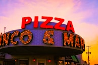 A retro-style sign prominently displays 'PIZZA' in bold red letters with a background of hexagonal patterns. Illuminated lights highlight the words on the marquee, creating an inviting and nostalgic atmosphere. The sky in the background displays a warm gradient of colors transitioning from orange to pink, suggesting a sunset scene.