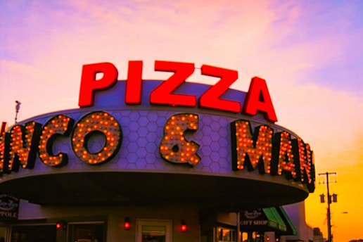 A retro-style sign prominently displays 'PIZZA' in bold red letters with a background of hexagonal patterns. Illuminated lights highlight the words on the marquee, creating an inviting and nostalgic atmosphere. The sky in the background displays a warm gradient of colors transitioning from orange to pink, suggesting a sunset scene.