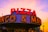 A retro-style sign prominently displays 'PIZZA' in bold red letters with a background of hexagonal patterns. Illuminated lights highlight the words on the marquee, creating an inviting and nostalgic atmosphere. The sky in the background displays a warm gradient of colors transitioning from orange to pink, suggesting a sunset scene.