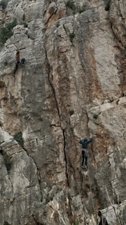 Two rock climbers are ascending a steep and rugged cliff face. The climber on the left is higher up, gripping the rock with hands and feet. The climber on the right is positioned lower and appears to be closely following a crack in the cliff. The scene conveys a sense of adventure and challenge against the backdrop of a natural and jagged rock formation.