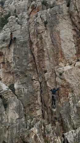 Two rock climbers are ascending a steep and rugged cliff face. The climber on the left is higher up, gripping the rock with hands and feet. The climber on the right is positioned lower and appears to be closely following a crack in the cliff. The scene conveys a sense of adventure and challenge against the backdrop of a natural and jagged rock formation.